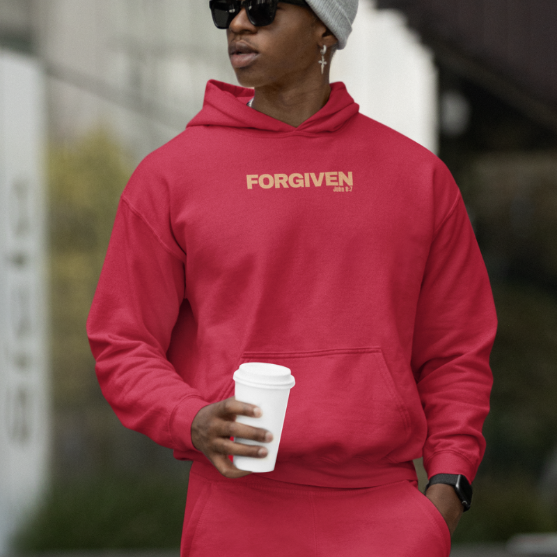 Man wearing a red hoodie and joggers set from the Forgiven Collection with dark shades a grey beanine and a gold dangling earring with a cross pendant gazing over his right shoulder quizzically